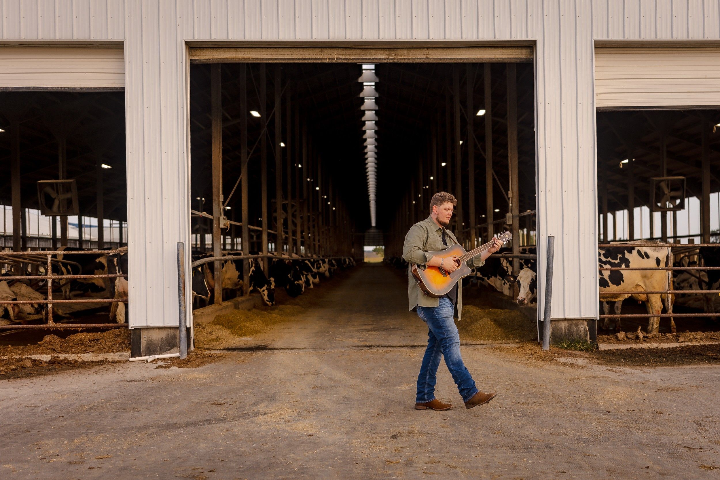 Isaac Buschur walking and playing acoustic guitar past an open dairy barn